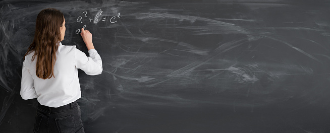 student woman or teacher in the class writes math formulas in chalk on a black chalkboard. young girl stands with her back to the camera. Back to school. Education concept. Banner. Place for your text