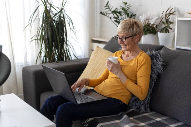 短いブロンドの髪を持つ年配の女性は、リビングルームのソファに座って、ビデオチャットを介してラップトップ上の友人とチャット - furniture internet adult blond hair ストックフォトと画像