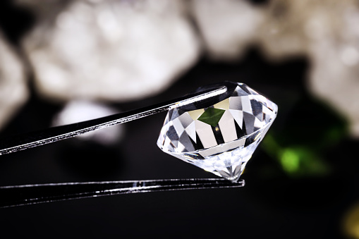 cut diamond held by a jeweler's tweezers, with rough diamond stones in the background, spot focus