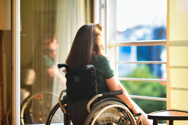Woman in wheelchair in front of open window in her apartment watching sunset stock photo