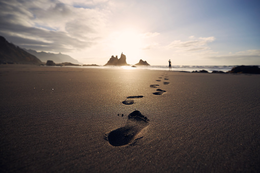 Footprints in sand against silhouette of person. Man walking along beach to sea at golden sunset. Tenerife, Canary Islands, Spain.
