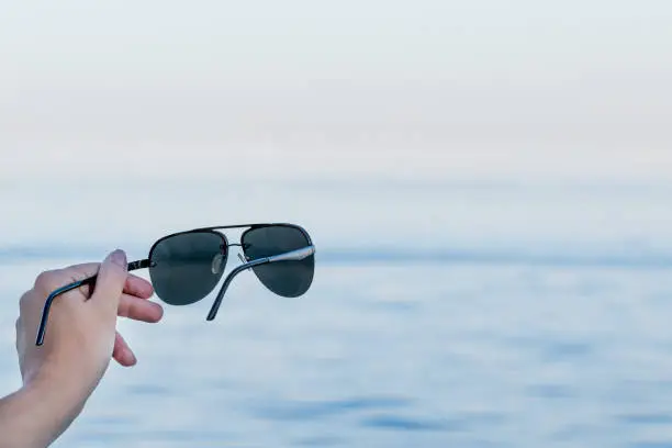 Photo of Sunglasses model aviators in a female hand on a blurred background of blue water on vacation at sea. Summer vacation concept with sunglasses and copy space