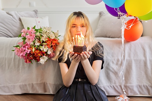 Birthday of female teenager, girl with birthday small cake with burning candles, room, bouquet of flowers, colored balloons background. Holiday, teens, age, celebration concept