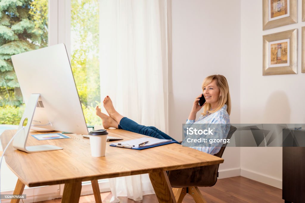 Attractive middle aged woman sitting at desk and working from home Shot of happy blond haired woman making a phone call and using computer while sitting at desk and working from home. Feet up on desk. Home office. Office Stock Photo