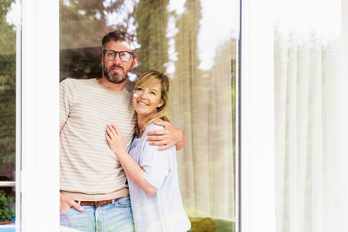 Through the glass shot of happy couple standing together at home while looking out the window.