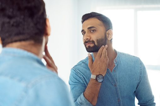Curious thoughtful indian man standing in front of mirror touching beard on face looking at reflection. Thoughtful young adult eastern guy with bristle engaged in morning facial skin care routine
