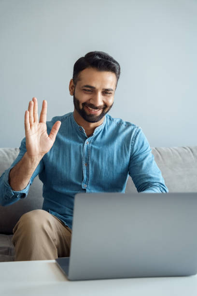 Portrait of indian business man waving hand to online interlocutor using laptop stock photo