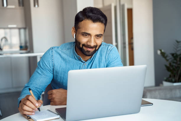 Portrait of young adult indian man wearing earphones looking at laptop screen stock photo