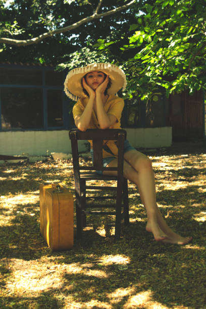 country woman sits on a wooden chair with a straw hat yellow shirt and jean shorts. - fashion women denim farm imagens e fotografias de stock