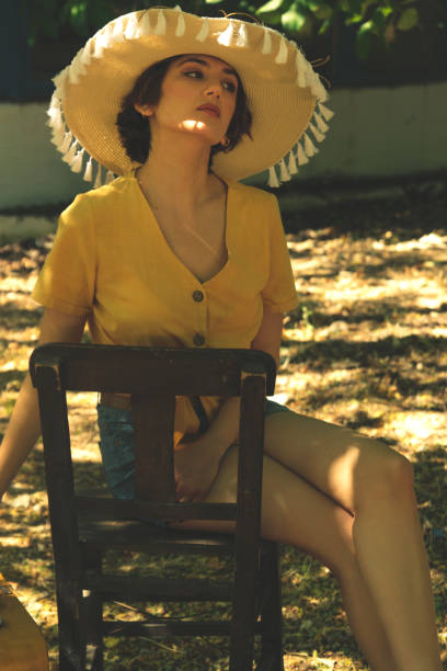 country woman sits on a wooden chair with a straw hat yellow shirt and jean shorts. - fashion women denim farm imagens e fotografias de stock