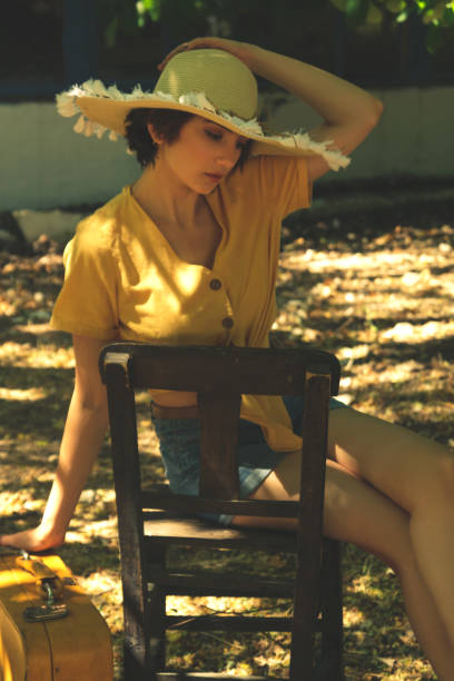 country woman sits on a wooden chair with a straw hat yellow shirt and jean shorts. - fashion women denim farm imagens e fotografias de stock