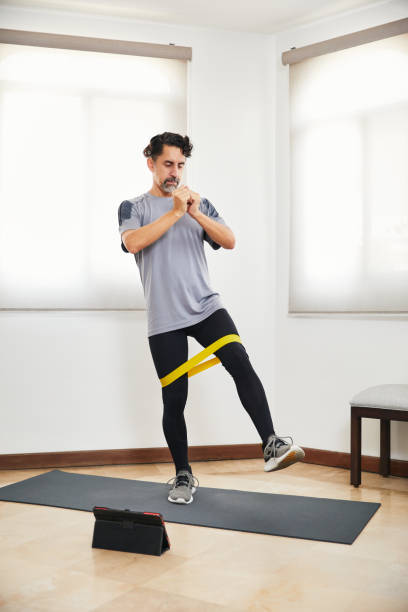 istock Middle aged man with beard doing exercise at home with rubber band looking video on tablet 1326998377