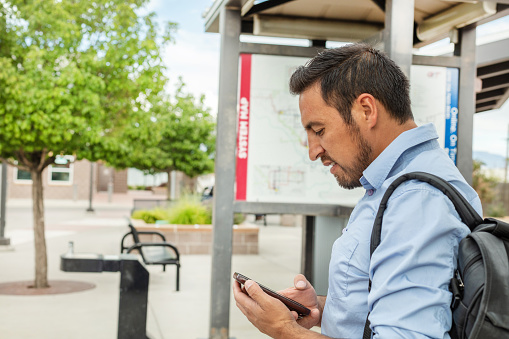 In Western Colorado Millennial Hispanic Male Commuter Waiting for Public Transportation at Bus Stop with Smartphone Technology Photo Series (Shot with Canon 5DS 50.6mp photos professionally retouched - Lightroom / Photoshop - original size 5792 x 8688 downsampled as needed for clarity and select focus used for dramatic effect)