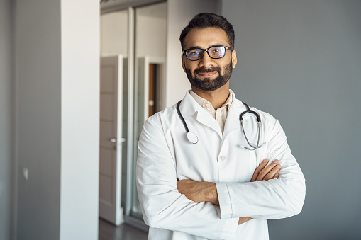 Portrait of male doctor wearing white lab coat, stethoscope standing and looking at camera in clinic hall. Arabian indian therapist, general practitioner headshot. Medicine, heal insurance, healthcare