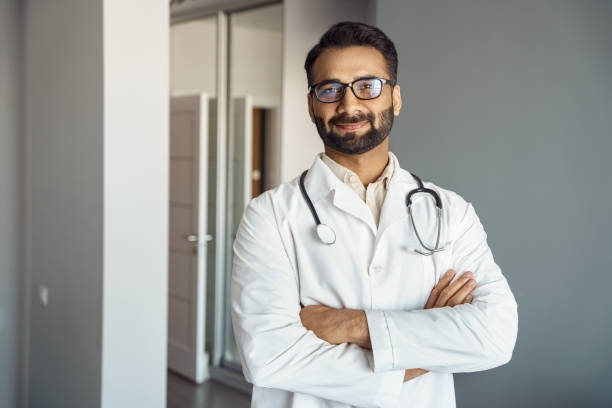 Portrait of male doctor in white coat and stethoscope standing in clinic hall Portrait of male doctor wearing white lab coat, stethoscope standing and looking at camera in clinic hall. Arabian indian therapist, general practitioner headshot. Medicine, heal insurance, healthcare laboratory coat stock pictures, royalty-free photos & images