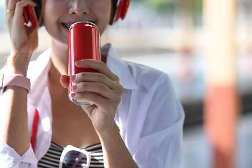 Smiling young female listening music in headphone and drinking sweet beverage from red can.
