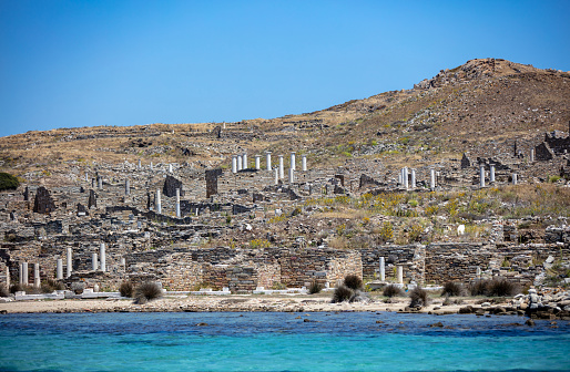 Delos Cyclades island, Greece. General view approaching by boat. Holy sanctuary archaeological site, UNESCO Heritage monument. Stone walls and marble pillars ruins, on rocky hill. Blue sky and sea