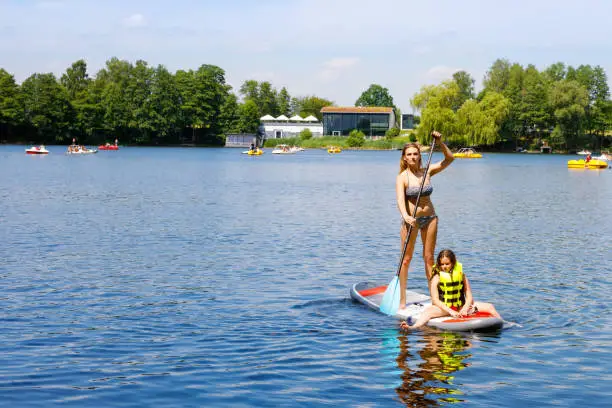 Young woman with school girl paddling on sup board on a lake. Active family on modern trendy stand up paddle board. Summer outdoors vacations activity for family and children. Watersport activieties