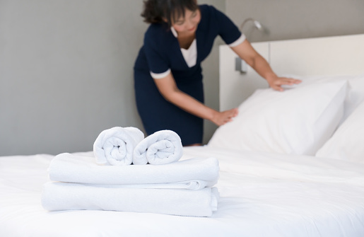 Clean white towels on bed with young hotel maid making bed on background