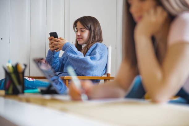 istock Teenage girl using a mobile phone, her sister doing homework 1327045363