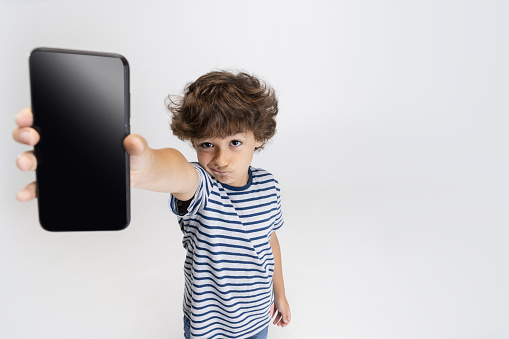 Demonstrating device screen. One Caucasian preschool boy in casual clothing with smartphone posing isolated on white studio background. Copyspace for ad