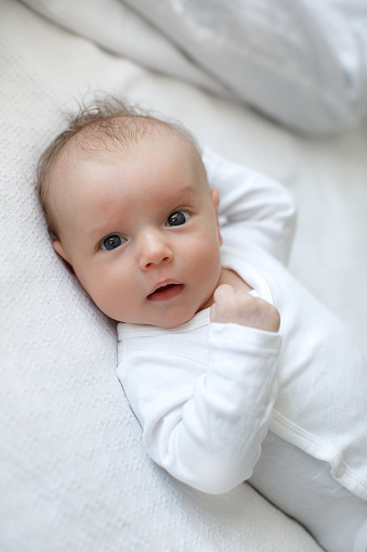 A charming boy in a white sunny bedroom plays alone, lying on the bed. A beautiful newborn baby with chubby cheeks lies on the bed, innocently looking at something, being naughty. Cute toddler playing alone. A newborn baby is having fun alone.