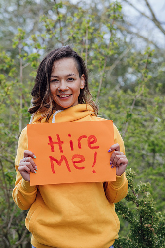 A young woman standing outside in front of trees, holding a piece of paper with 'hire me' on it. She is looking at the camera and smiling.