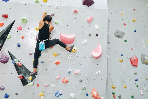 Fit athlete climbing up bouldering wall without auto belay devices, view from the back