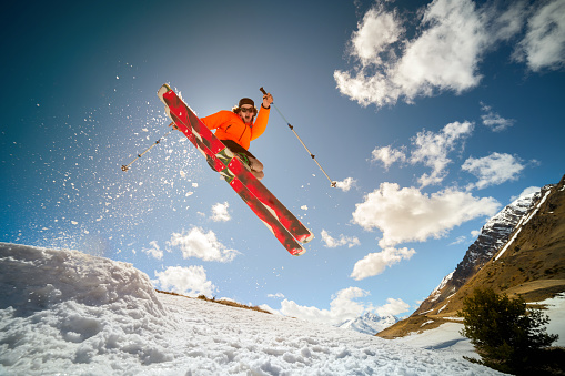 Beautiful young caucasian man in an orange jacket and black shorts jumping from a springboard on red skis against the background of mountains, sky, clouds, sun