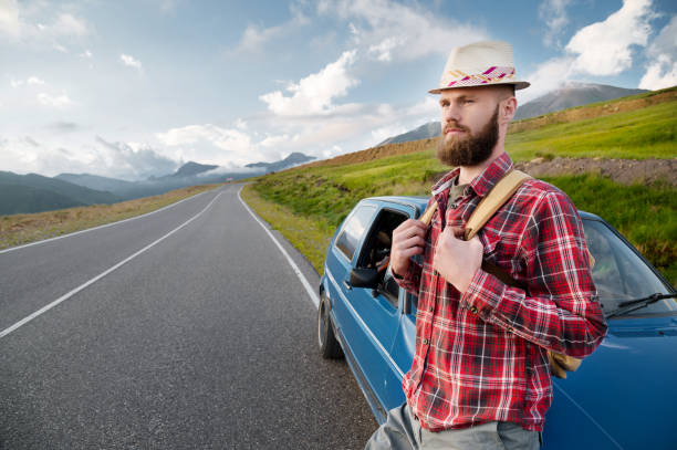 istock Attractive bearded man in a plaid red shirt and hat with a backpack. Stands next to a retro car on the background of a picturesque road in the mountains at sunset. Easy travel, hitchhiking 1327063912