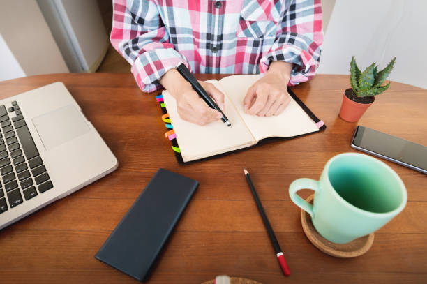 istock Young Caucasian attractive woman taking notes in her notebook while sitting in the kitchen at the table. Paper notebook writing female hands 1327064068
