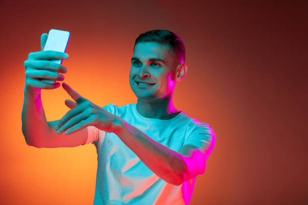 istock Portrait of young Caucasian man using phone isolated over gradient red orange studio background in neon light with copyspace for ad. Concept of human emotions 1327067865