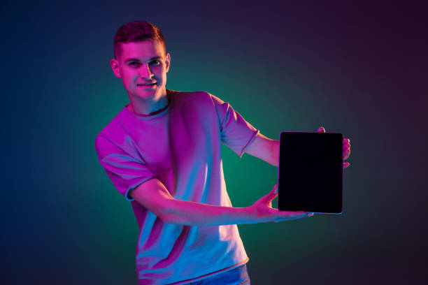 istock Young Caucasian man with digital tablet isolated over gradient blue green studio background in pink neon light with copyspace for ad. Concept of human emotions 1327067887
