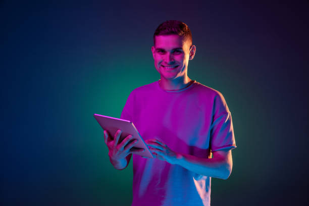 istock Young Caucasian man with digital tablet isolated over gradient blue green studio background in pink neon light with copyspace for ad. Concept of human emotions 1327067890