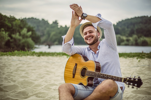 One young man, a handsome young man playing guitar on the beach. A smiling guy playing an acoustic guitar. A carefree man looking to the side of the seashore.