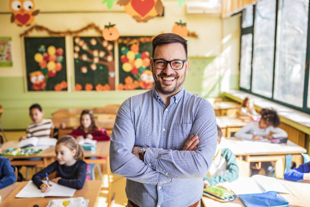 istock Happy elementary teacher with crossed arms in the classroom. 1327072498