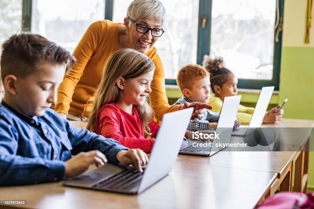 Happy mature teacher assisting her students on computer class at school. Happy mature teacher helping her students in using computers on a class at elementary school. Focus is on happy girl. Child Stock Photo