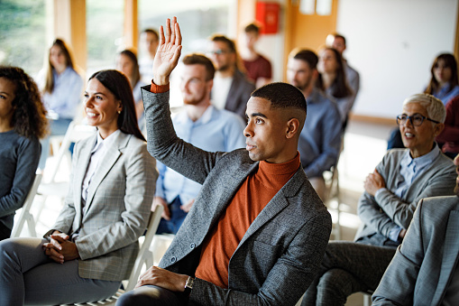 African American businessman attending a seminar with his colleagues while raising his hand to ask a question.