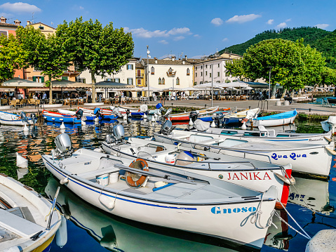 Garda,Verona,Italy 01 June 2021:Little harbor of Garda with moored boats, the tables of the restaurants along the edges of the lake and people strolling along the lake