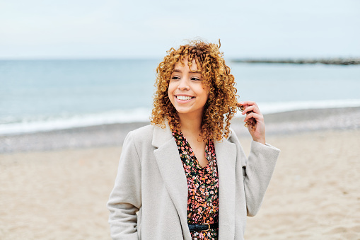 Portrait of an Afro-American woman with curly hair walking along the shore of the beach on a cloudy day.