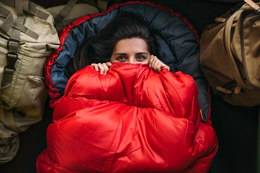 A young woman in a comfortable sleeping bag in a tent, top view. A tourist in a sleeping bag. A traveler wrapped in a red sleeping bag. Travel, camping concept, adventure. Traveling with a tent