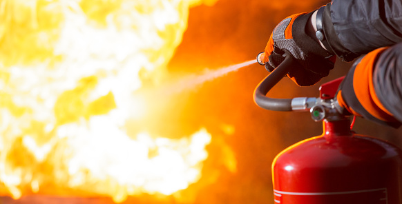 Close-up showing a person pointing the nozzle of a fire extinguisher at the base of a large fire.
