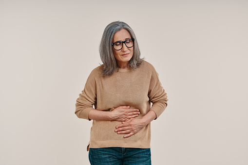 Frustrated mature woman suffering from stomachache while standing against grey background