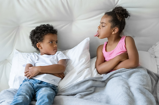 African American sister and brother quarreling. Preteen girl showing tongue to younger boy. Two little children lying in bed closeup portrait. Bad mood, negative emotion, upbringing and family concept