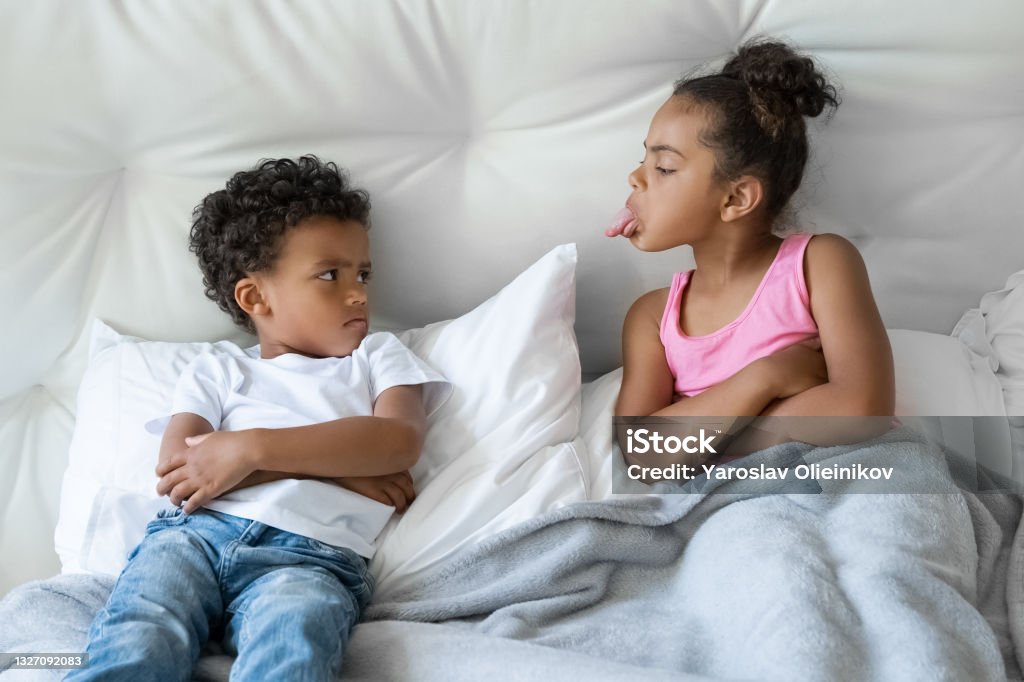 African American sister and brother little children quarreling lying in bed African American sister and brother quarreling. Preteen girl showing tongue to younger boy. Two little children lying in bed closeup portrait. Bad mood, negative emotion, upbringing and family concept Sibling Stock Photo