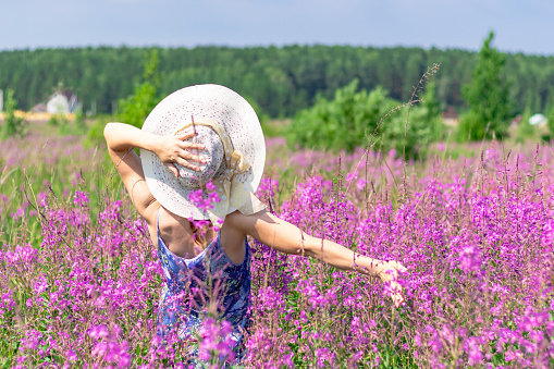 Young woman in a field of flowers in a straw hat against a background of green forest and blue sky on a hot summer day. Selective focus