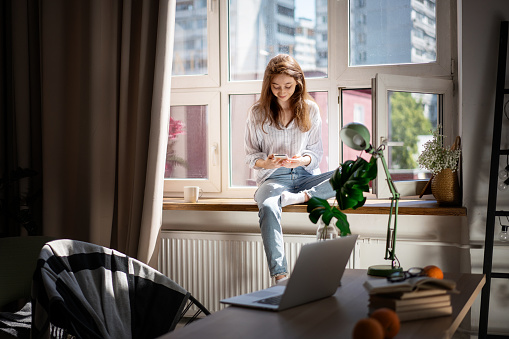 Beautiful happy young woman using smartphone sitting by the window in a modern apartment