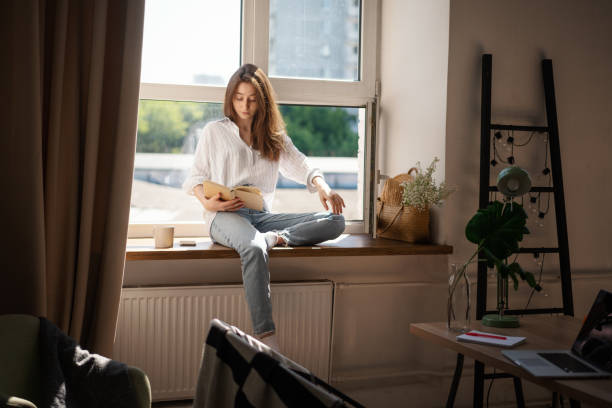 istock Young beautiful woman student reading book at home while sitting on window in modern apartment 1327095151