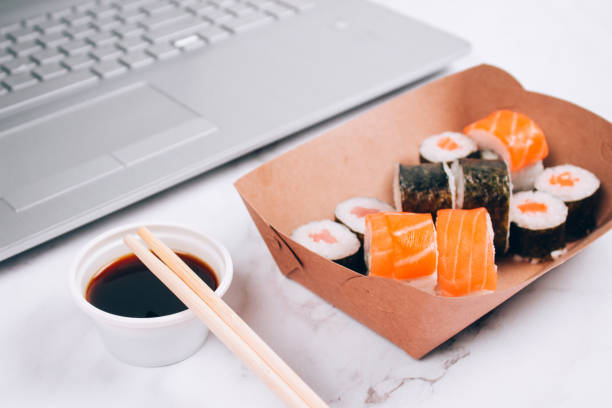 istock Different sushi rolls with salmon in delivery paper box on white marble table background with laptop - futomaki, uramaki and hosomaki. Online order food and delivery food concept 1327096365
