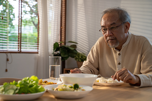 Unhappy Asian Senior older man sit alone, eat foods on table in house. Depressed mature attractive elderly retired grandfather stay at home with painful face feeling upset, lonely and miss his family.